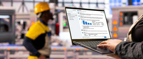 Gestor em ambiente industrial observa informações em um notebook na operação, com profissionais ao fundo usando equipamentos de segurança, ilustrando monitoramento e controle do trabalho.