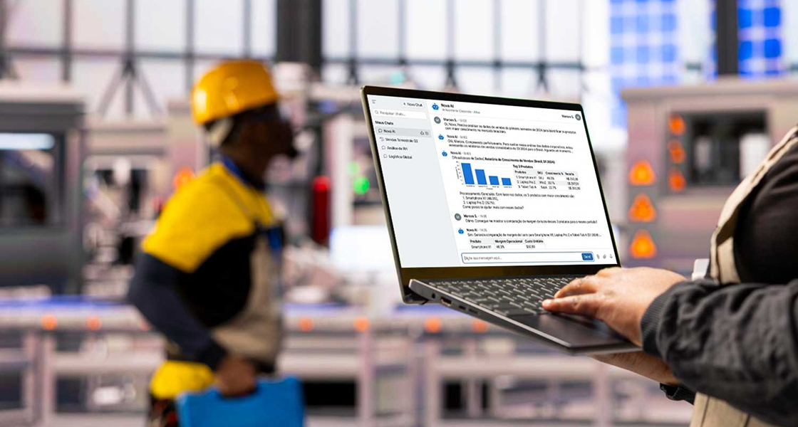 Gestor em ambiente industrial observa informações em um notebook na operação, com profissionais ao fundo usando equipamentos de segurança, ilustrando monitoramento e controle do trabalho.