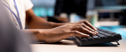 Foto nas mãos de uma pessoa digitando em um teclado preto sobre uma mesa de escritório. Ao fundo, telas de computador aparecem desfocadas em um ambiente corporativo corporativo de TI ou contabilidade. A imagem representa o trabalho de atualização de dados, cadastro de fornecedores e validação de nota fiscal em um sistema de gestão empresarial (ERP industrial), simbolizando a adequação tecnológica necessária nas indústrias para mitigar os impactos e riscos operacionais com a chegada do CNPJ alfanumérico e as exigências de créditos da Reforma Tributária.