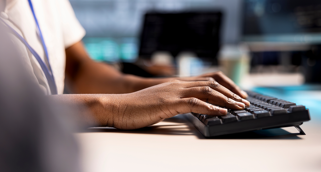 Foto nas mãos de uma pessoa digitando em um teclado preto sobre uma mesa de escritório. Ao fundo, telas de computador aparecem desfocadas em um ambiente corporativo corporativo de TI ou contabilidade. A imagem representa o trabalho de atualização de dados, cadastro de fornecedores e validação de nota fiscal em um sistema de gestão empresarial (ERP industrial), simbolizando a adequação tecnológica necessária nas indústrias para mitigar os impactos e riscos operacionais com a chegada do CNPJ alfanumérico e as exigências de créditos da Reforma Tributária.