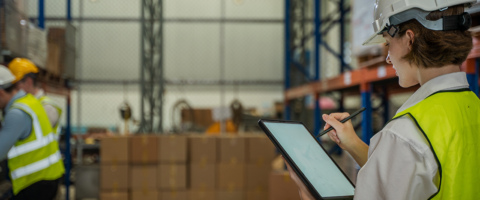 Trabalhadores em centro de armazenamento e logística, com capacete e colete de segurança, usando o sistema de gestão empresarial (ERP) no tablet para acompanhar a operação na indústria